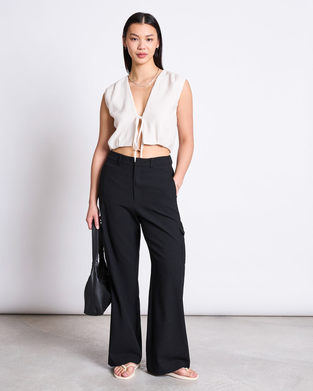 A woman with straight, dark hair and a neutral expression stands against a plain background wearing jan-n-junes CARGO PANTS FANO BLACK—high-waisted black cargo pants made from recycled polyester—paired with a sleeveless cream tie-front top, sandals, and a black bag.
