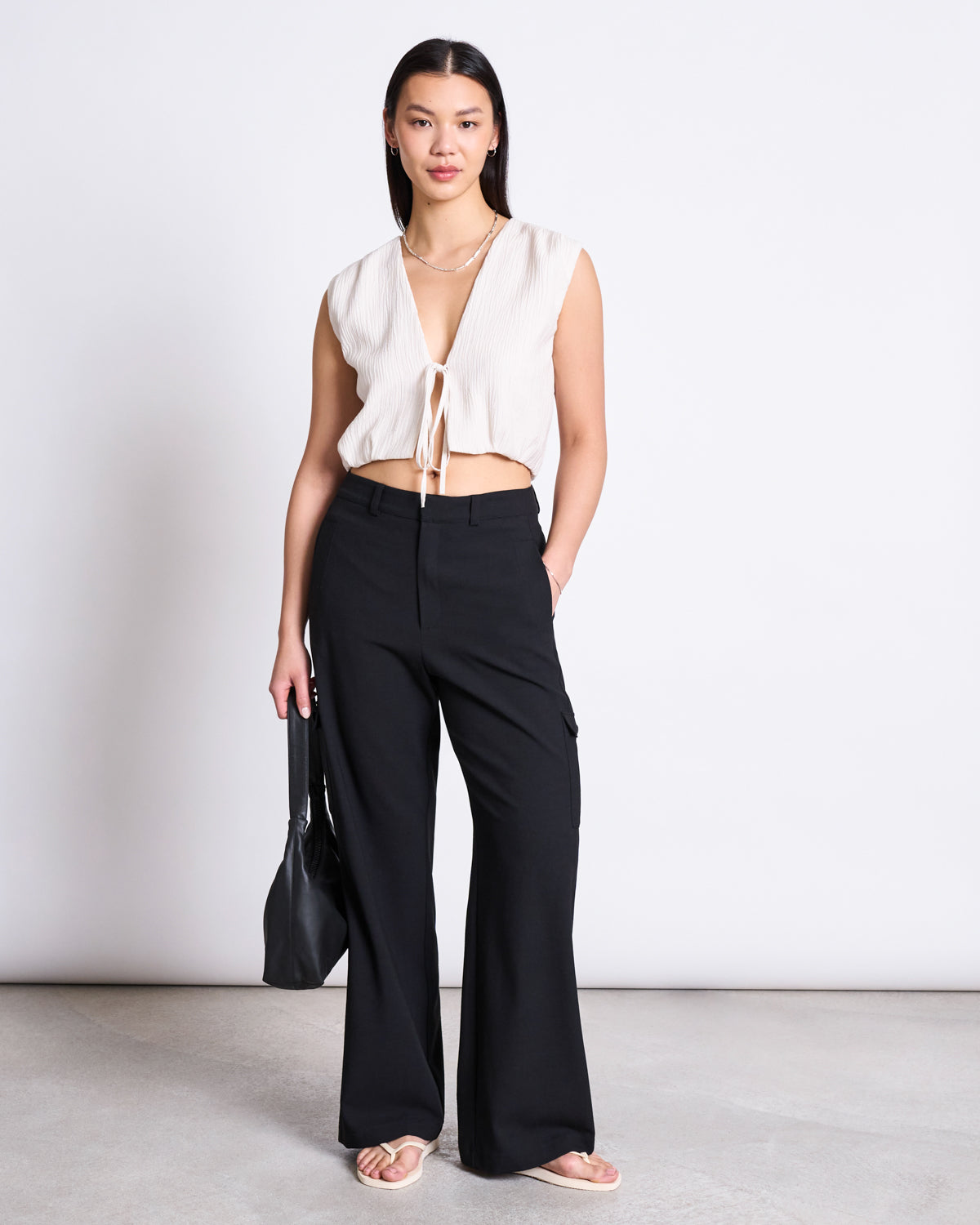 A woman with straight, dark hair and a neutral expression stands against a plain background wearing jan-n-junes CARGO PANTS FANO BLACK—high-waisted black cargo pants made from recycled polyester—paired with a sleeveless cream tie-front top, sandals, and a black bag.