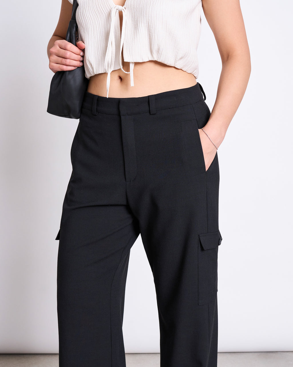A person wears jan-n-june’s CARGO PANTS FANO BLACK—high-waisted black cargo pants with pockets—paired with a short white tie-front crop top and holding a black bag. The photo is cropped from mid-torso to knees.