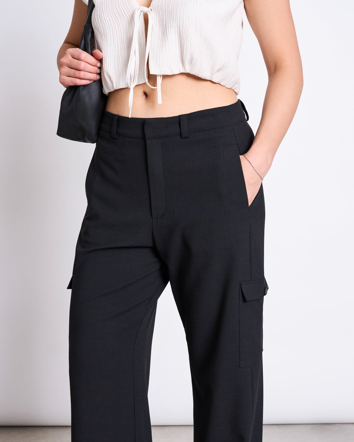 A person wears jan-n-june’s CARGO PANTS FANO BLACK—high-waisted black cargo pants with pockets—paired with a short white tie-front crop top and holding a black bag. The photo is cropped from mid-torso to knees.