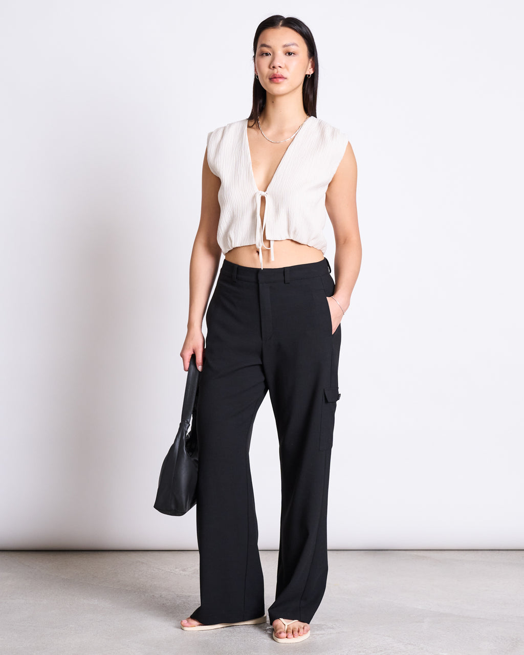 A woman stands against a plain background wearing jan-n-junes CARGO PANTS FANO BLACK, a sleeveless white tie-front crop top, and sandals. She holds a black bag and has one hand in her pocket.