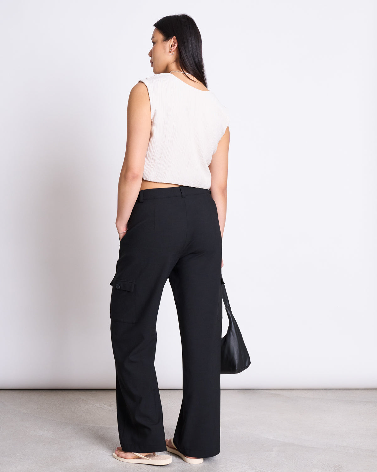 A woman faces away in jan-n-june’s CARGO PANTS FANO BLACK, paired with a sleeveless white top and sandals. She holds a black bag in her left hand against a plain white background.