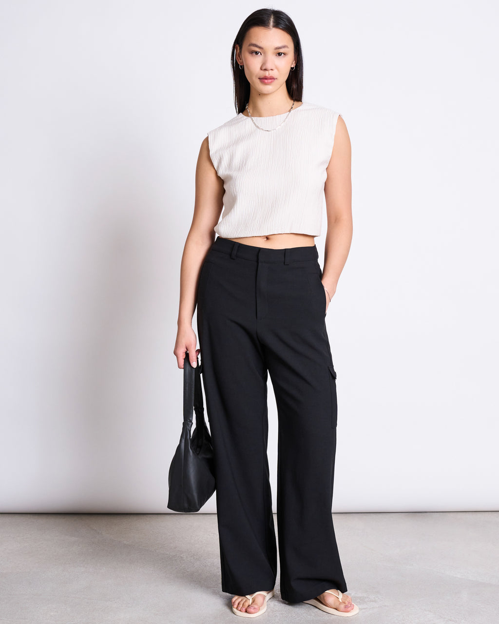 A woman with straight, shoulder-length hair stands against a plain background wearing jan-n-junes CARGO PANTS FANO BLACK, a sleeveless white top, sandals, and holding a black bag.