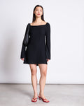A woman with straight dark hair and a neutral expression stands against a plain background, wearing the jan-n-june SQUARE NECK MINI DRESS GLAD BLACK with long sleeves, red heeled sandals, a black shoulder bag, and a delicate necklace.