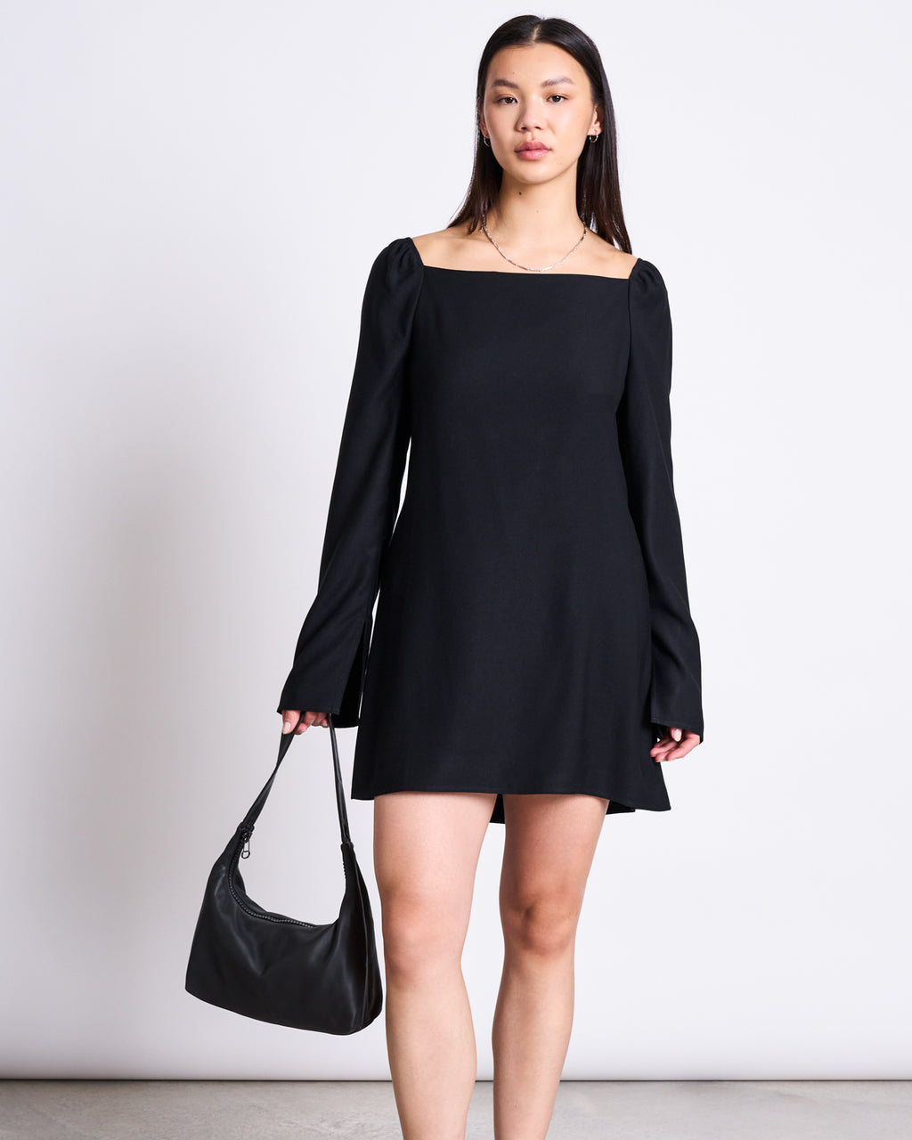 A woman with straight hair and simple jewelry stands against a white background wearing the jan-n-june SQUARE NECK MINI DRESS GLAD BLACK with long sleeves, holding a black handbag, and embodying timeless elegance.