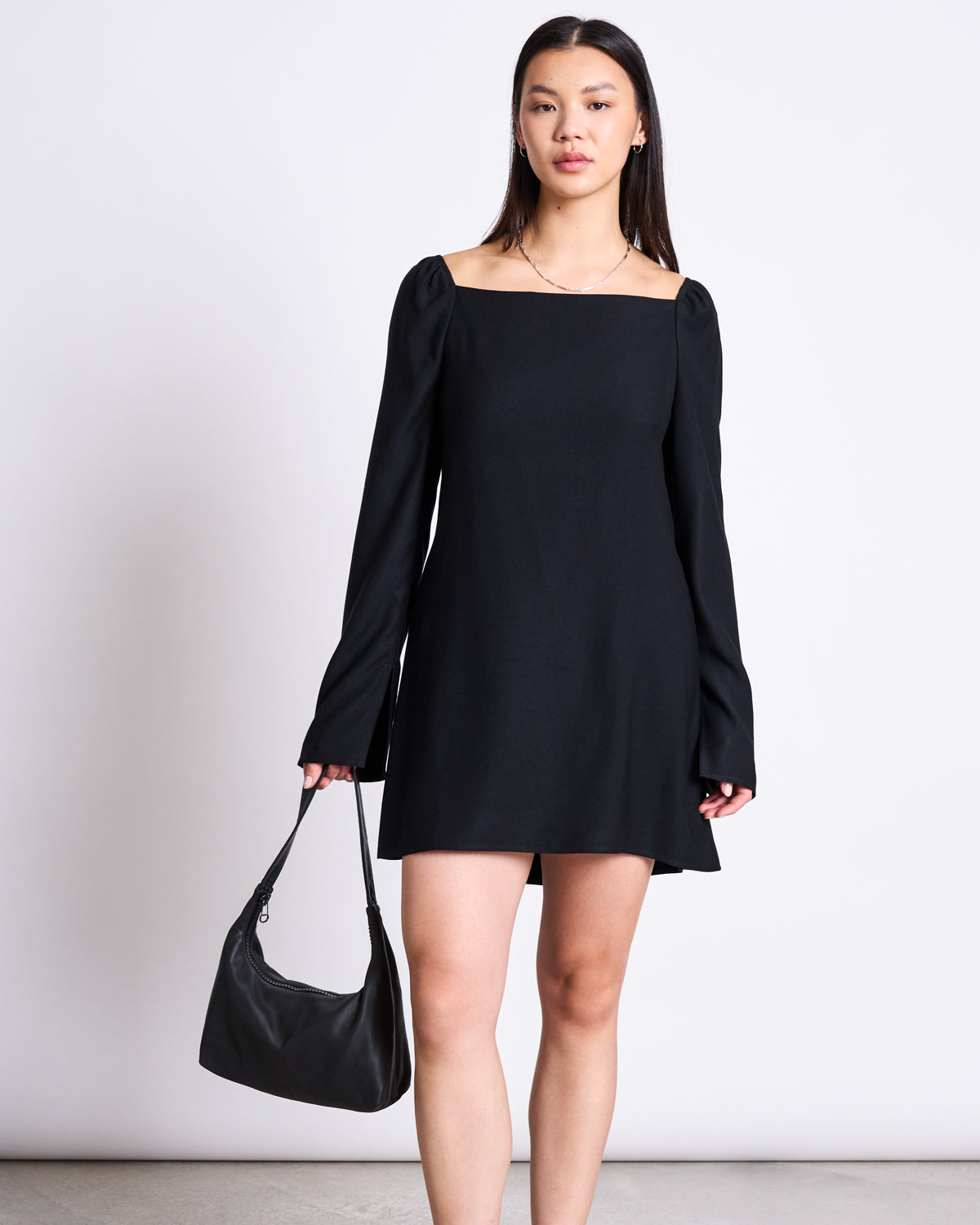 A woman with straight hair and simple jewelry stands against a white background wearing the jan-n-june SQUARE NECK MINI DRESS GLAD BLACK with long sleeves, holding a black handbag, and embodying timeless elegance.