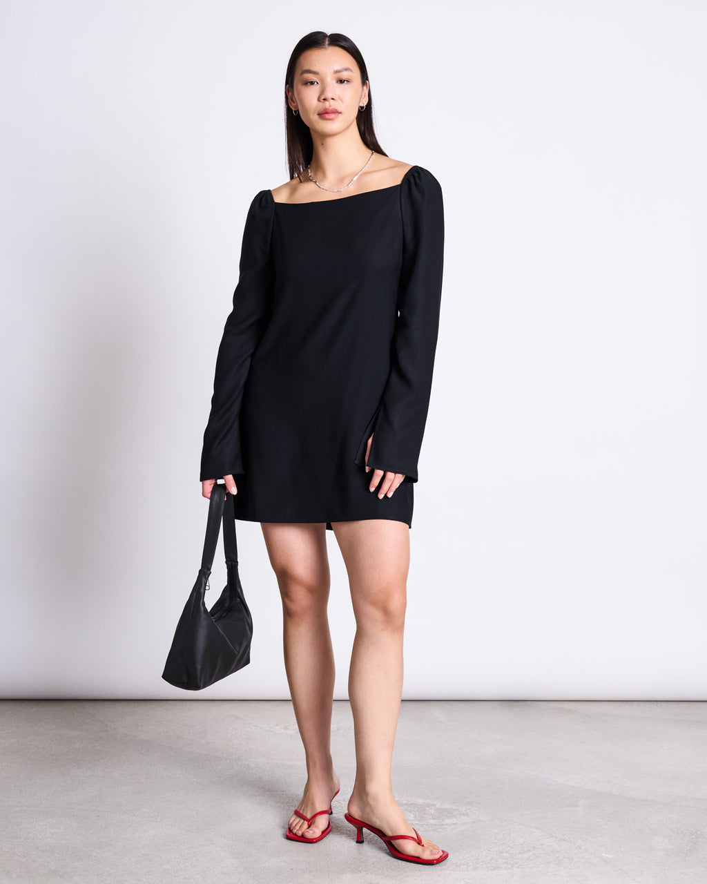A woman with long dark hair and a neutral expression stands against a plain background wearing the jan-n-june SQUARE NECK MINI DRESS GLAD BLACK, red heeled sandals, and holding a black handbag.