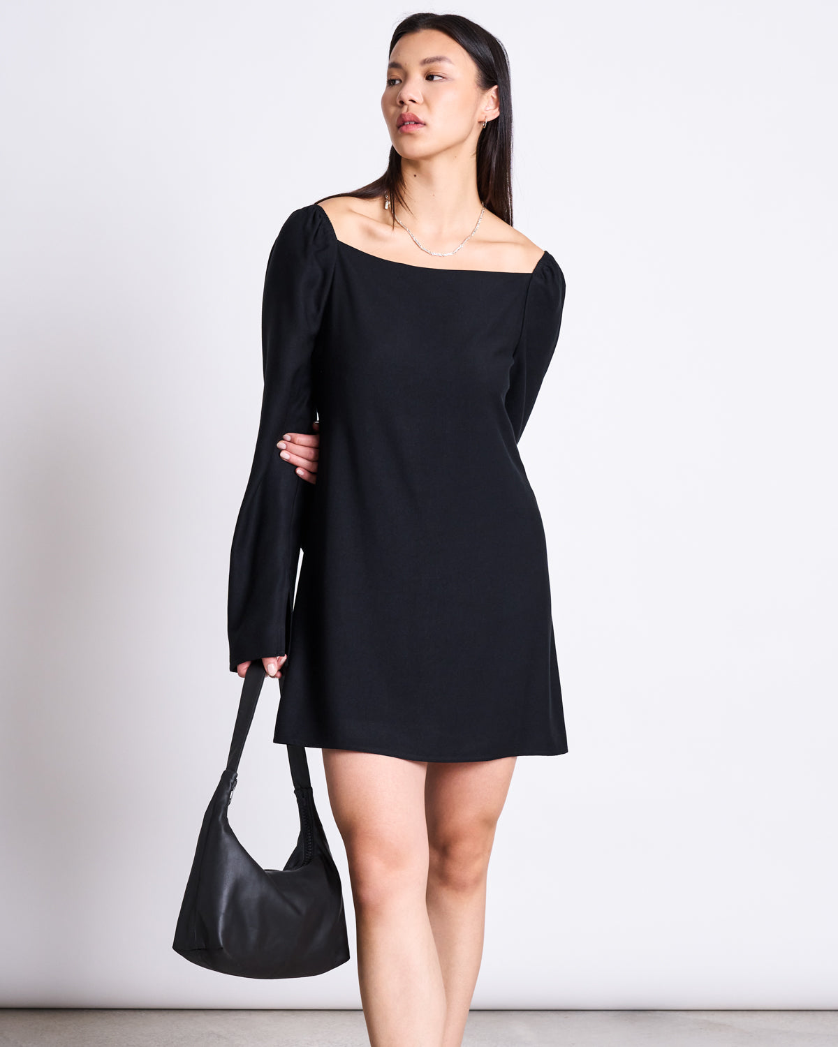 A woman wears the jan-n-june SQUARE NECK MINI DRESS GLAD BLACK with long sleeves, holding a black handbag at her side. She stands against a plain white background, gazing slightly to the side.