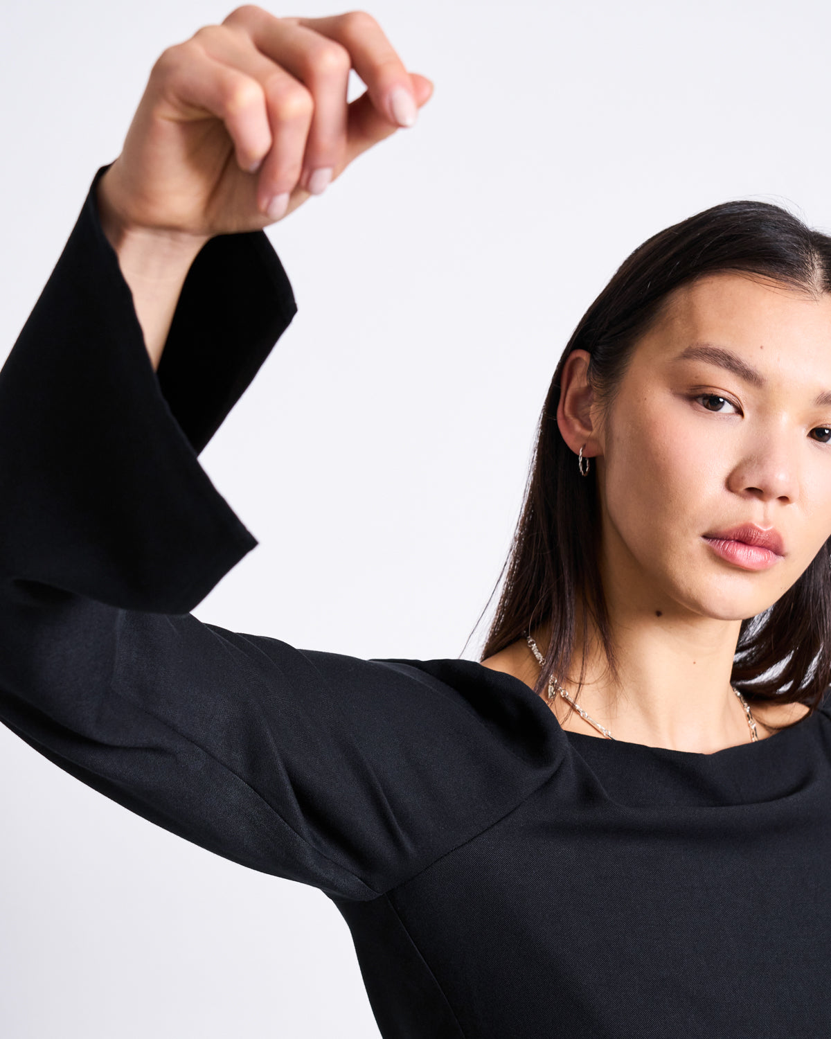 A woman with straight dark hair poses in the jan-n-june SQUARE NECK MINI DRESS GLAD BLACK, featuring flared sleeves and a square neckline. She wears small hoop earrings and natural makeup against a plain white background.