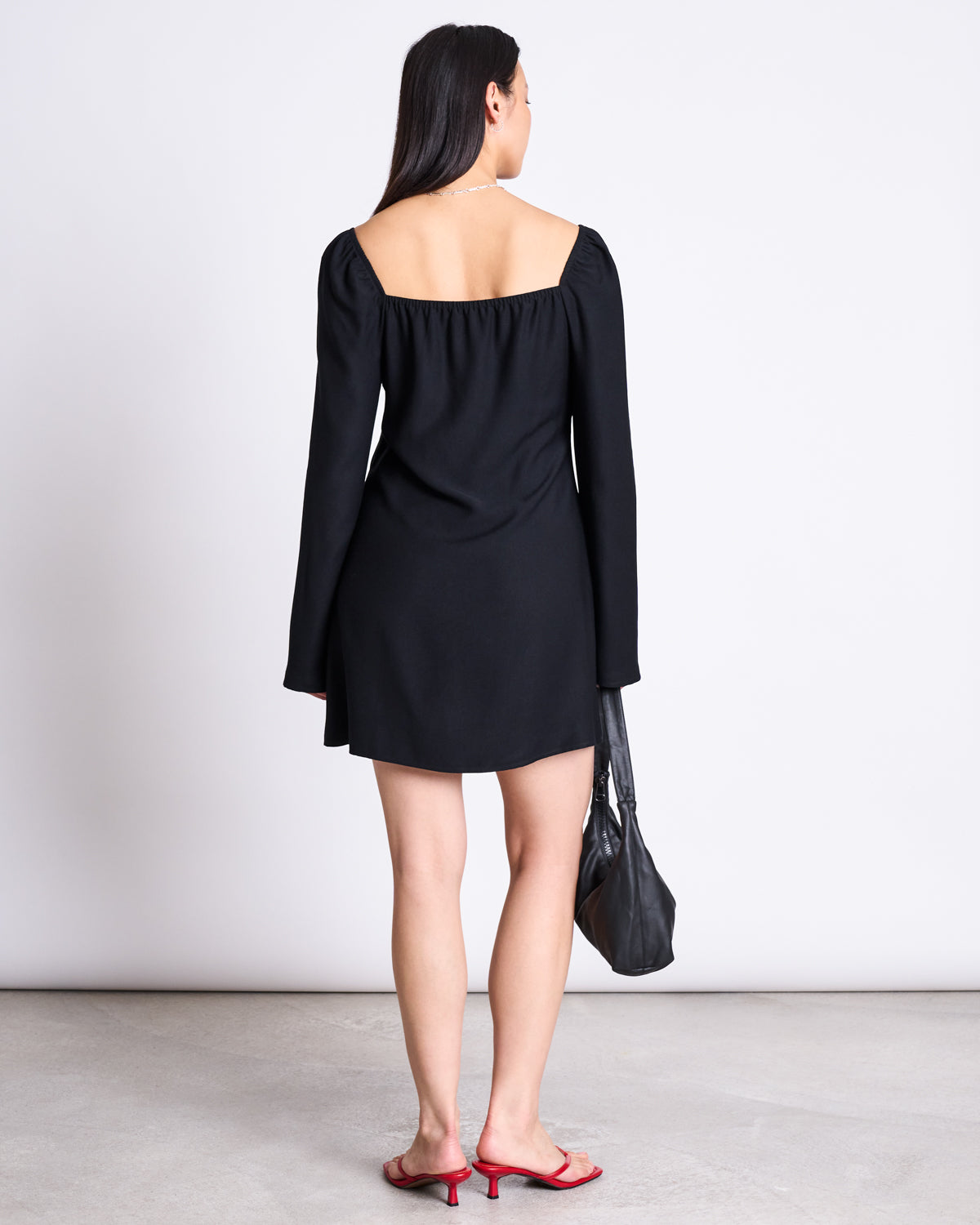 A woman stands facing away, wearing the jan-n-june SQUARE NECK MINI DRESS GLAD BLACK and red heeled sandals. She holds a black handbag by her side against a plain white background.