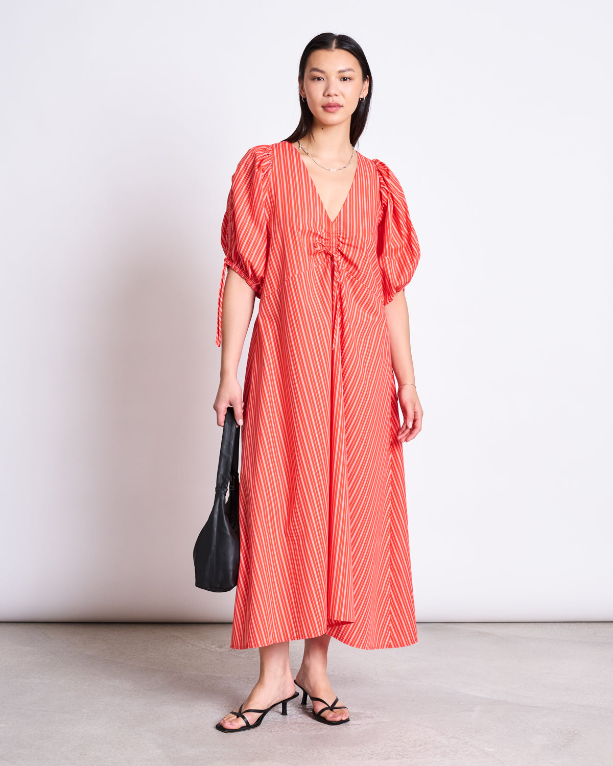 A woman with straight dark hair and a neutral expression models the jan-n-june MAXI DRESS DANS CHERRY RED WHITE STRIPED, featuring puffed sleeves and a v-neck, paired with black strappy sandals and a black handbag against a plain background.