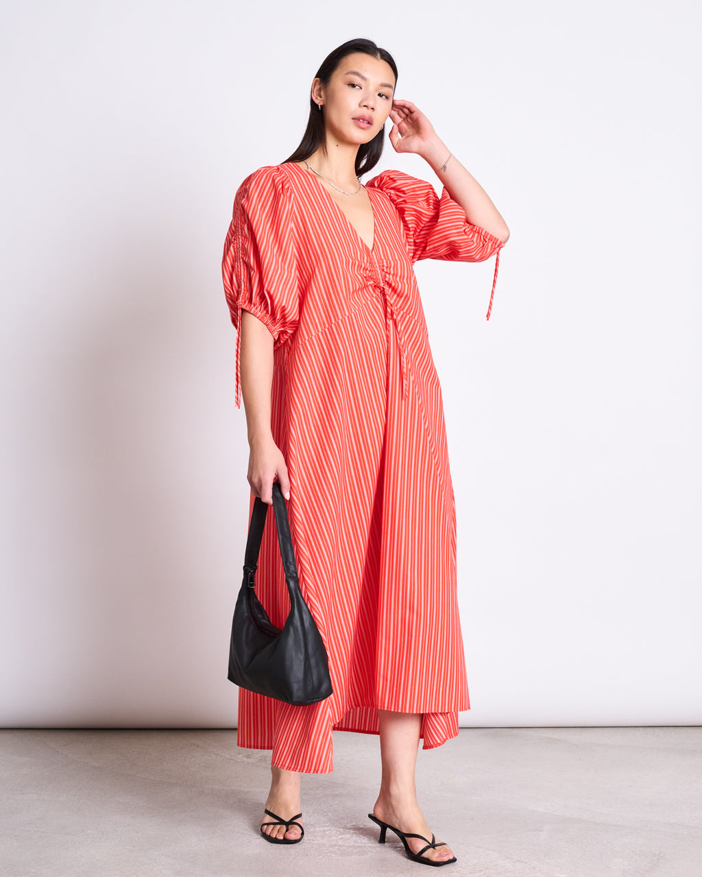 A woman stands against a plain background wearing the jan-n-june MAXI DRESS DANS CHERRY RED WHITE STRIPED with a v-neck and short puff sleeves. She holds a black handbag, wears black heeled sandals, and her straight hair is loose.