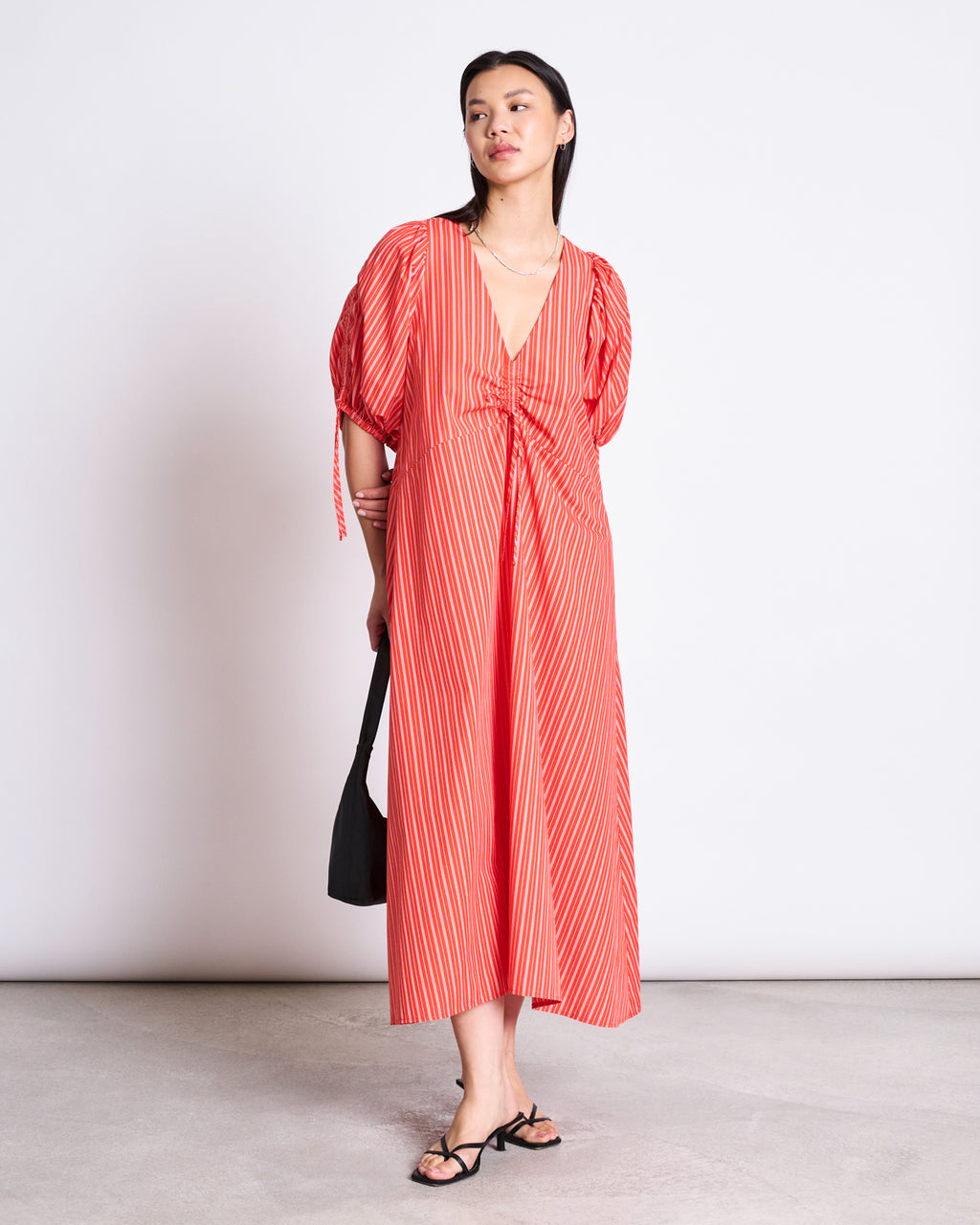 A woman stands against a plain background wearing the jan-n-june MAXI DRESS DANS CHERRY RED WHITE STRIPED, featuring organic cotton, puffed sleeves, and a deep V-neck. She pairs it with a black handbag and black sandals.