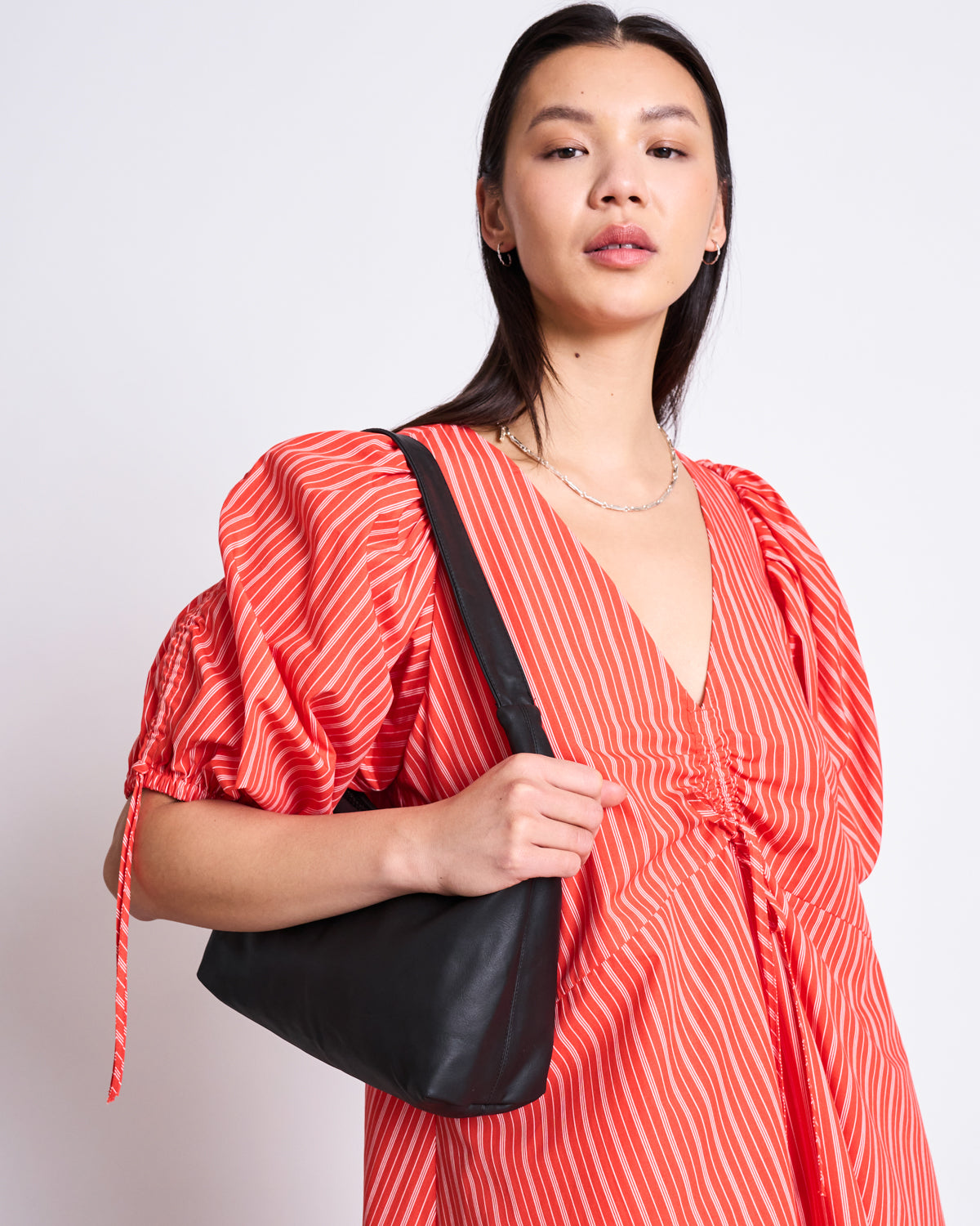 A woman with straight dark hair poses against a white background, wearing the jan-n-june MAXI DRESS DANS CHERRY RED WHITE STRIPED with puffy sleeves and a v-neck, paired with a black shoulder bag.