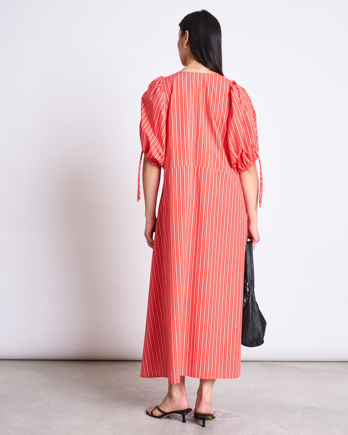 A woman with long, straight dark hair faces away, wearing the jan-n-june MAXI DRESS DANS CHERRY RED WHITE STRIPED with puffed sleeves, paired with black sandals and a black handbag.