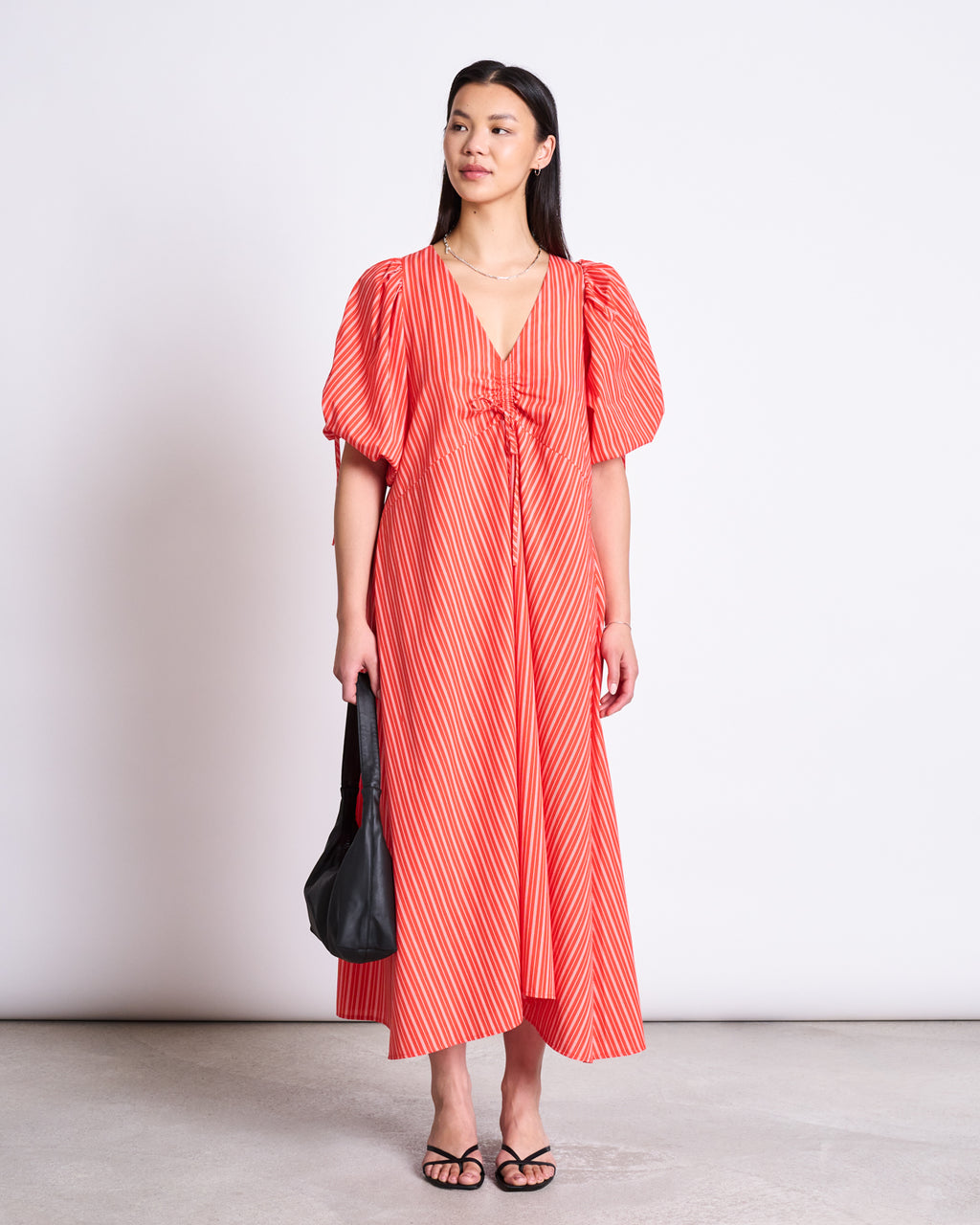 A woman in the jan-n-june MAXI DRESS DANS CHERRY RED WHITE STRIPED, a v-neck, puff-sleeve organic cotton maxi dress, stands against a plain background holding a black handbag and wearing black sandals.