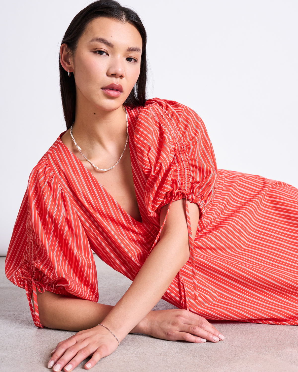 A woman with straight dark hair wears the jan-n-june MAXI DRESS DANS CHERRY RED WHITE STRIPED, featuring a v-neck and short puff sleeves. She sits on the floor, leaning on one arm, looking at the camera against a plain light background.
