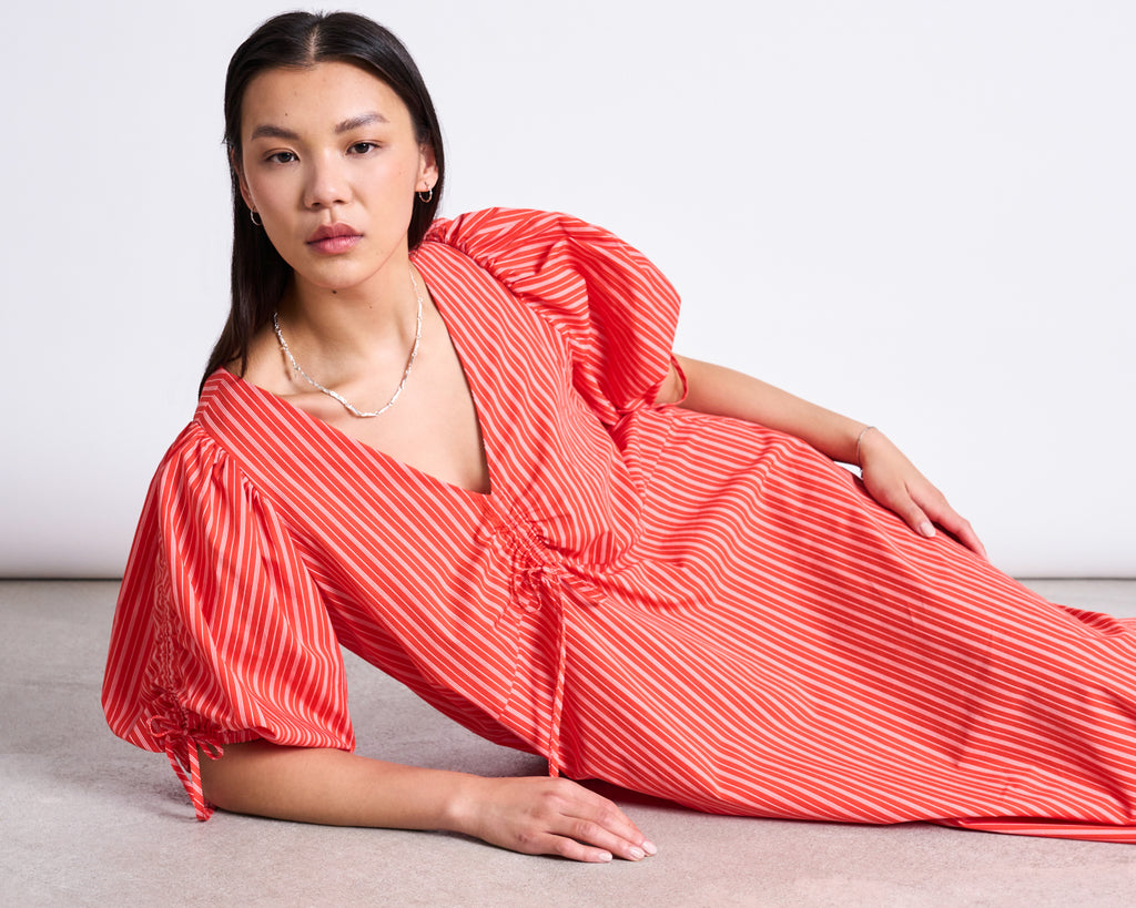 A woman with long dark hair reclines on the floor, wearing the jan-n-june MAXI DRESS DANS CHERRY RED WHITE STRIPED featuring organic cotton, puffed sleeves, and a V-neckline. She looks calmly at the camera against a light background.