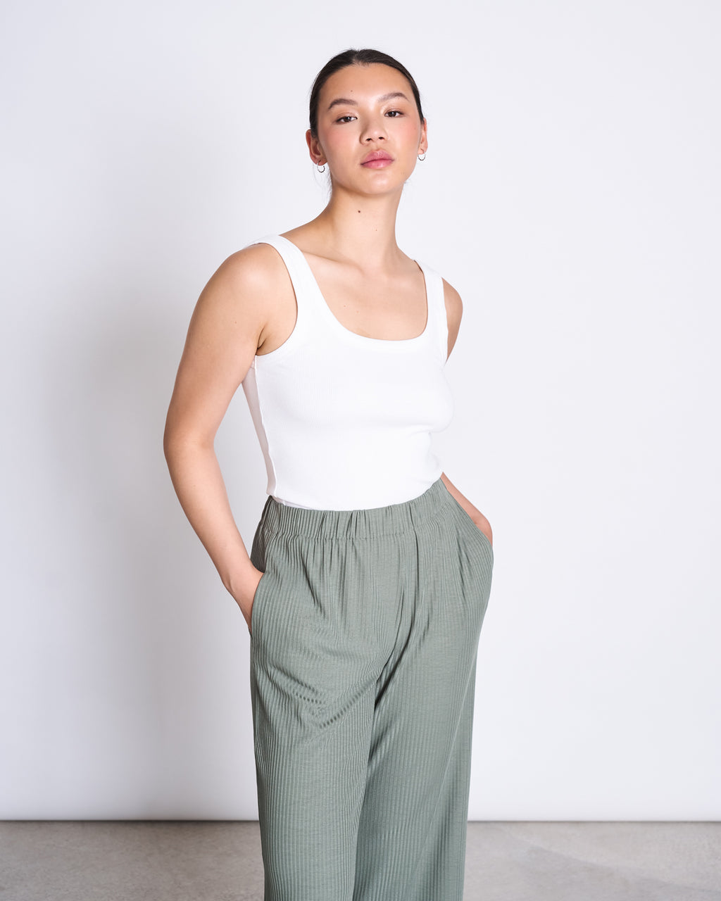 A person stands against a white background, wearing a sleeveless white top and jan-n-junes LYOCELL RIB PANTS SMILLA SEA SPRAY in sage green eco-friendly Tencel™ Lyocell, hands in pockets, hair pulled back with a neutral expression.