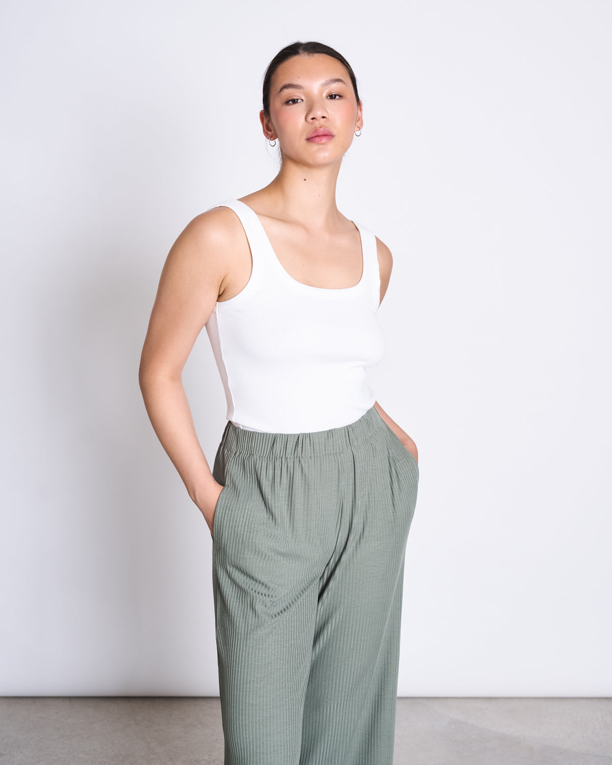 A person stands against a white background, wearing a sleeveless white top and jan-n-junes LYOCELL RIB PANTS SMILLA SEA SPRAY in sage green eco-friendly Tencel™ Lyocell, hands in pockets, hair pulled back with a neutral expression.