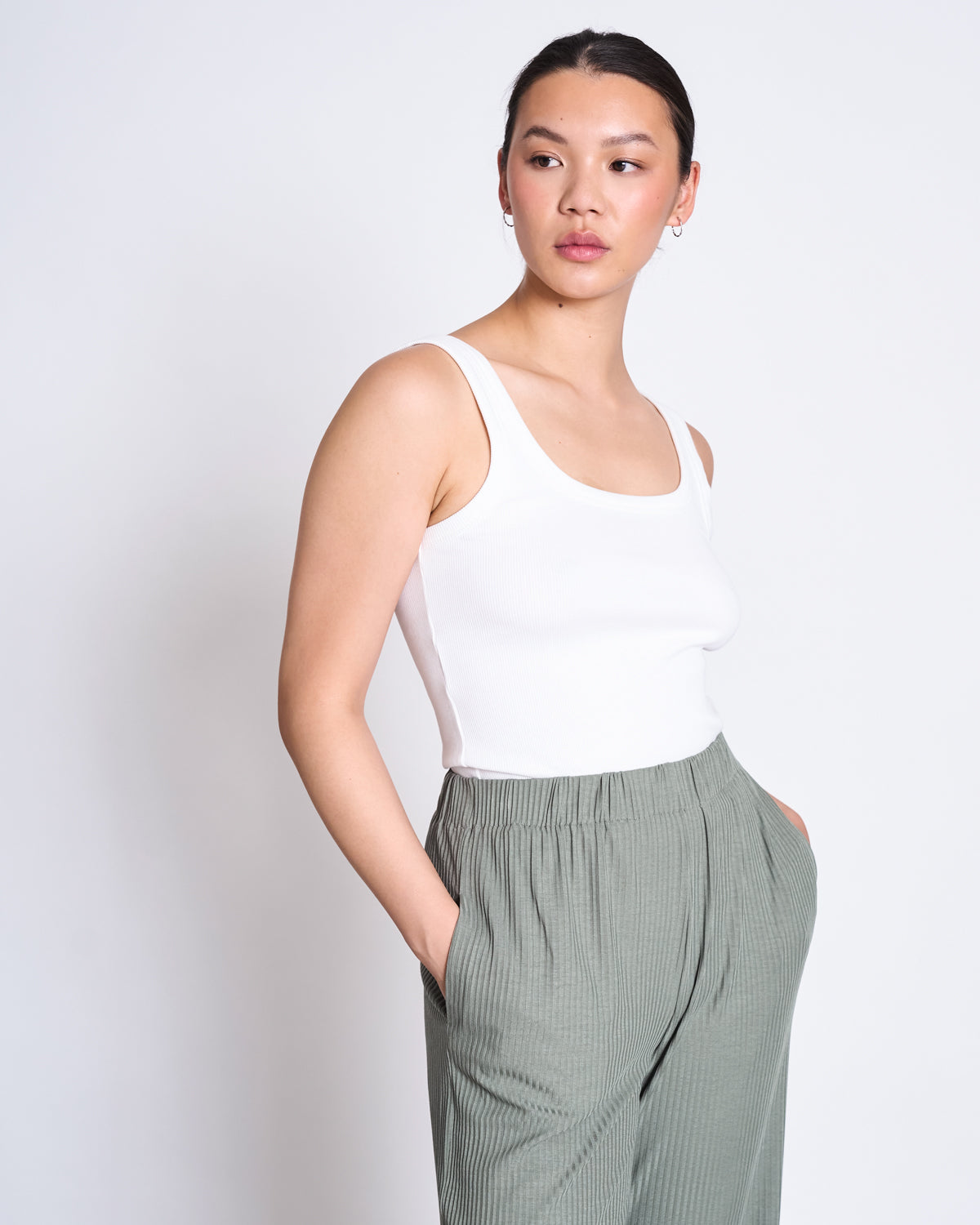 A woman stands against a plain background, wearing jan-n-junes LYOCELL RIB PANTS SMILLA SEA SPRAY—high-waisted, light green pleated pants made from eco-friendly Tencel™ Lyocell with an elastic waistband—as she looks to the side, hands in her pockets.