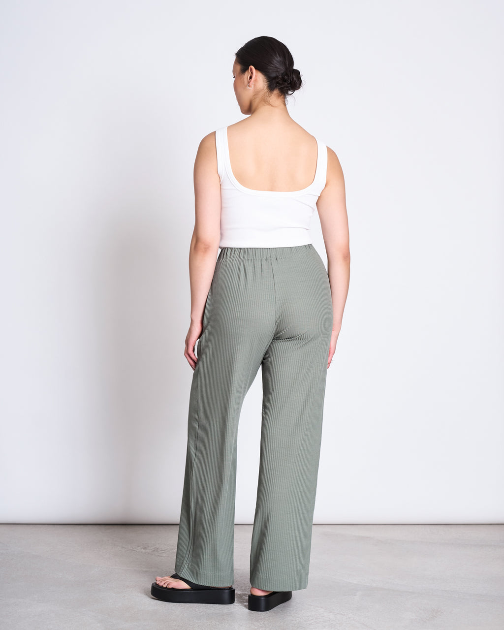 A woman faces away, wearing jan-n-junes LYOCELL RIB PANTS SMILLA SEA SPRAY in loose sage green Tencel™ Lyocell with an elastic waistband and a white tank top, paired with black sandals, against a plain white background.