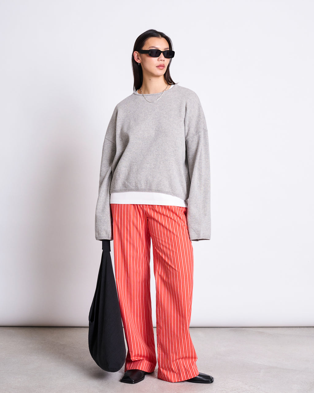 A person wearing jan-n-junes COTTON PANTS SOBY CHERRY RED WHITE STRIPED GOTS stands against a plain white background, styled with a loose gray sweater, black shoes, sunglasses, and a large black bag.