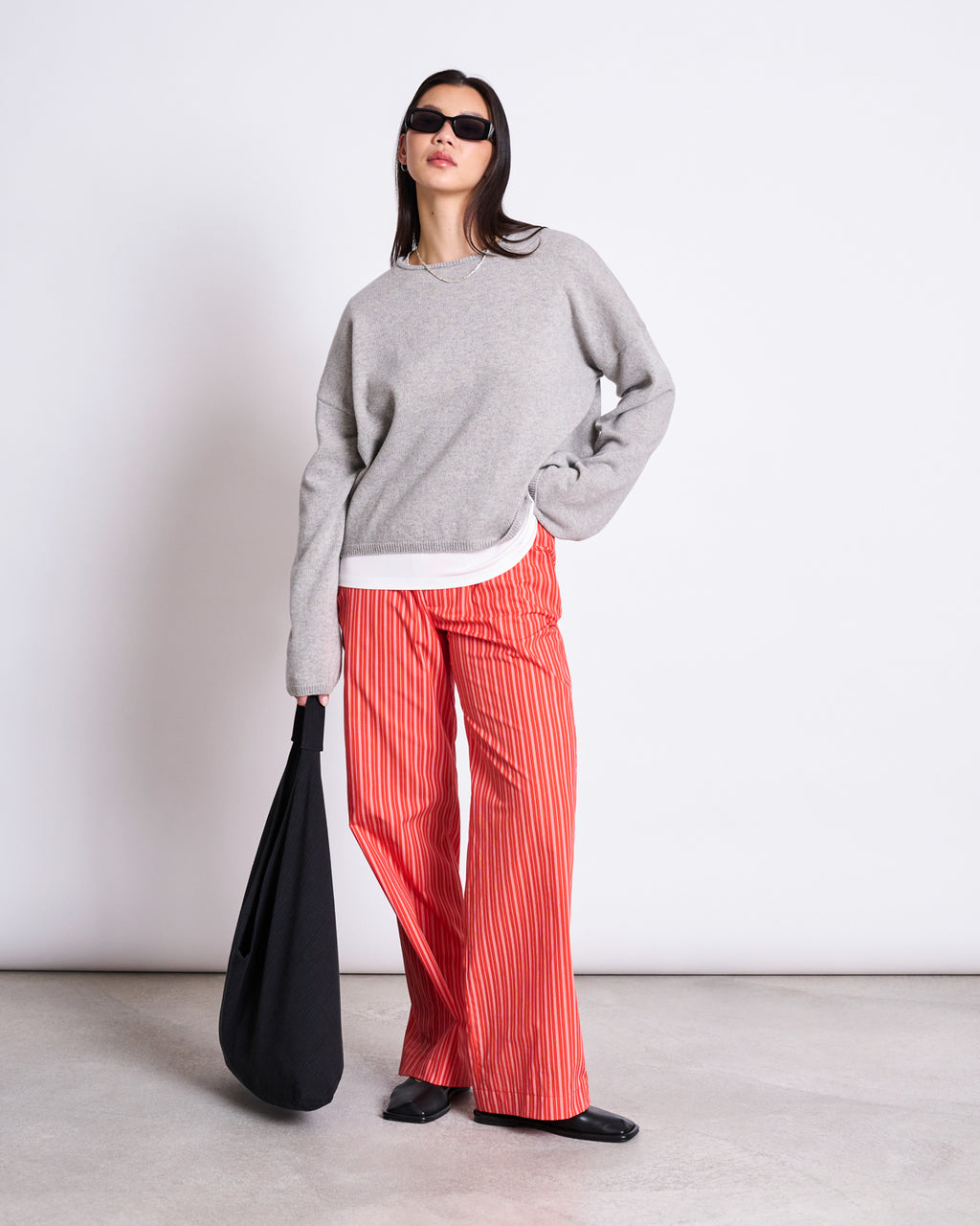 A woman poses against a plain background, wearing jan-n-junes COTTON PANTS SOBY CHERRY RED WHITE STRIPED GOTS, paired with a grey sweater, black shoes, sunglasses, and holding a large black bag.