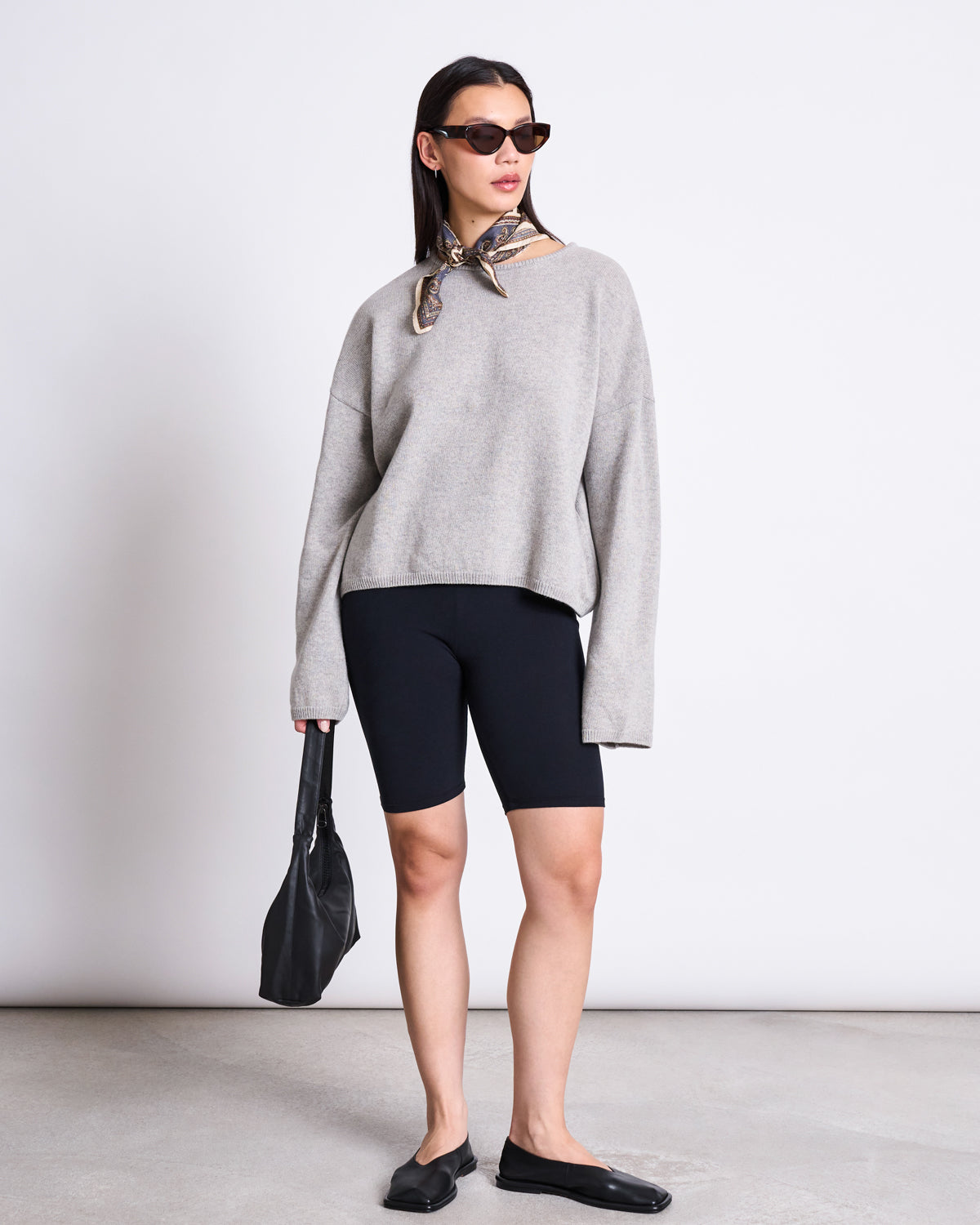 A woman stands against a plain background wearing black sunglasses, a gray oversized sweater, jan-n-june BIKER SHORTS BLACK GOTS, black flats, and a scarf around her neck. She holds a black handbag at her side.