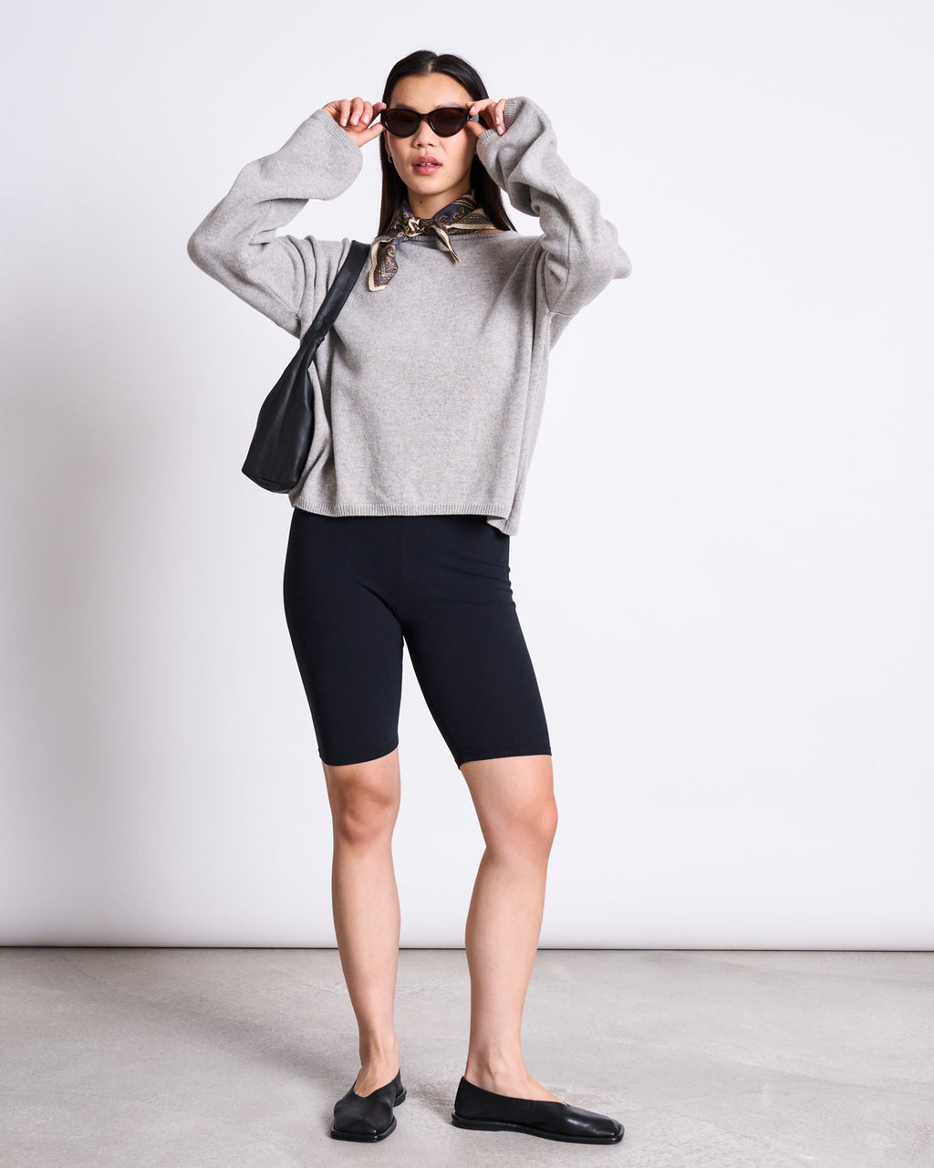 A woman stands against a plain background wearing jan-n-june’s BIKER SHORTS BLACK GOTS, a loose gray sweater, black flats, sunglasses, and a black shoulder bag, holding her sunglasses with both hands.