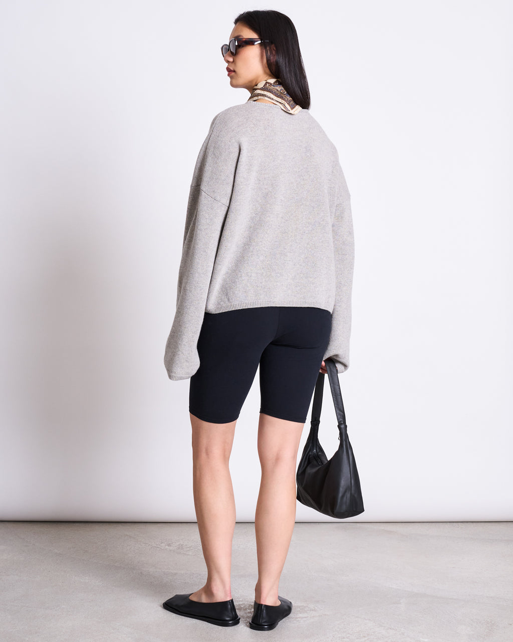 A woman faces away, wearing jan-n-junes BIKER SHORTS BLACK GOTS with an elastic waistband, a loose grey organic cotton sweater, slip-on shoes, sunglasses, a patterned scarf, and holding a black handbag against a light background.