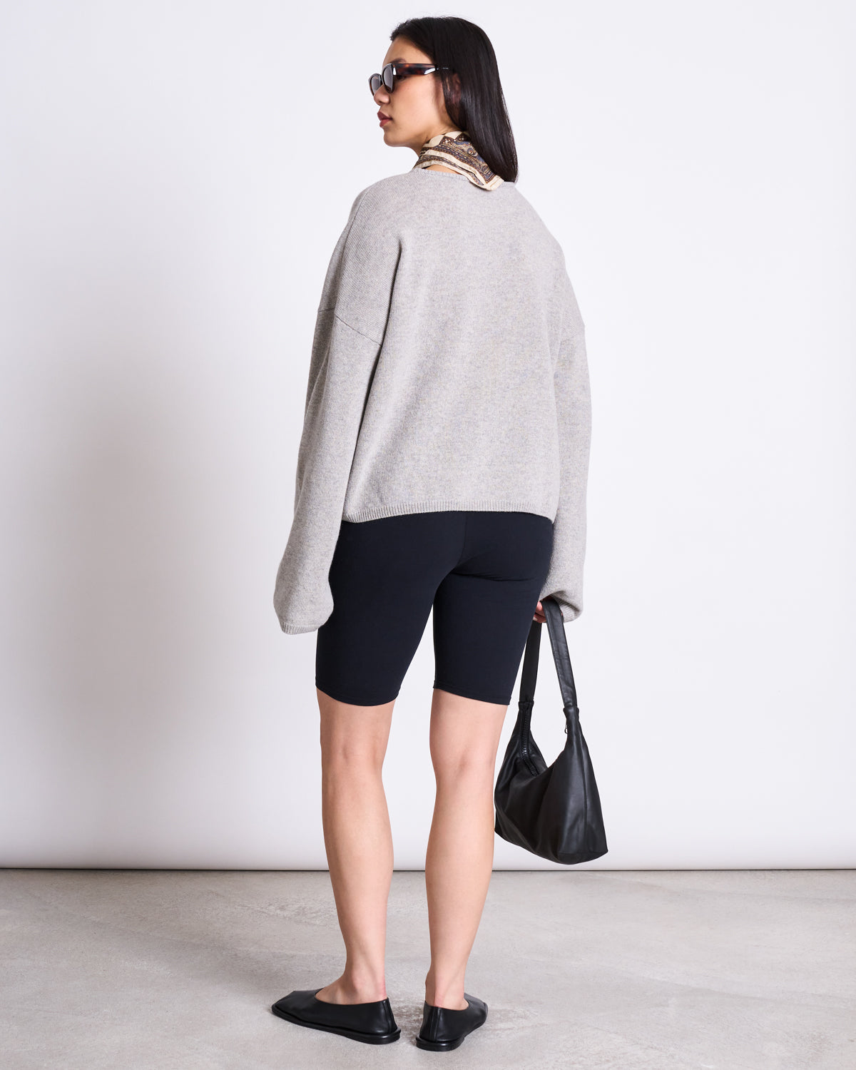 A woman faces away, wearing jan-n-junes BIKER SHORTS BLACK GOTS with an elastic waistband, a loose grey organic cotton sweater, slip-on shoes, sunglasses, a patterned scarf, and holding a black handbag against a light background.