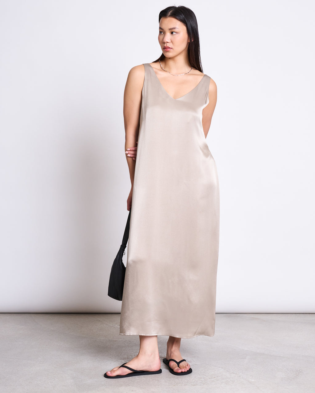 A woman stands against a plain background in the jan-n-june MAXI DRESS BENVIE SILVER GREY, a chic, sleeveless vegan silk alternative with a V-neckline, paired with black flip-flops and a black bag; her long hair is down as she gazes to the side.