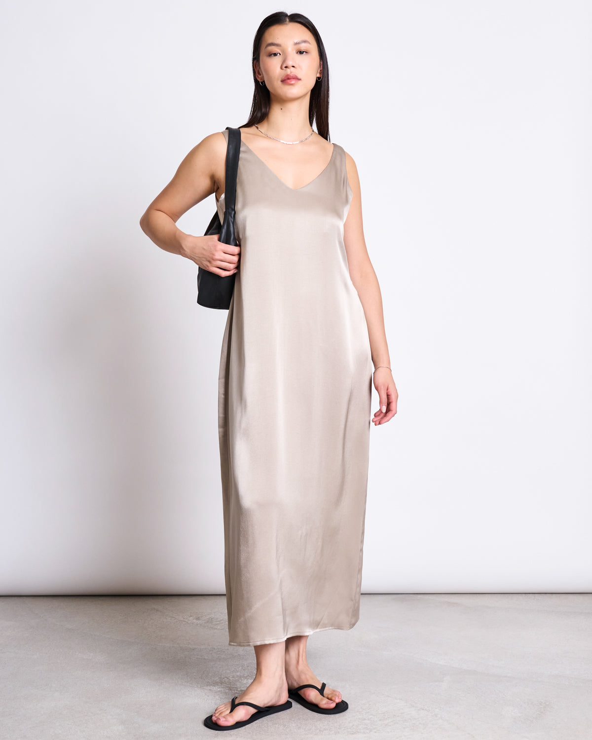 A woman stands against a plain background wearing the jan-n-june MAXI DRESS BENVIE in silver grey, styled with black sandals and a black shoulder bag. She has long straight hair and a relaxed expression.