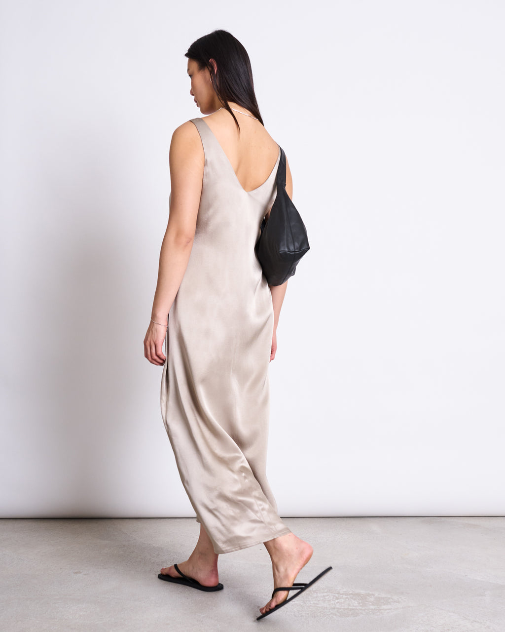 A woman with straight dark hair walks away from the camera in the jan-n-june MAXI DRESS BENVIE SILVER GREY, made from a vegan silk alternative, paired with black sandals and a black shoulder bag. The background is plain white.