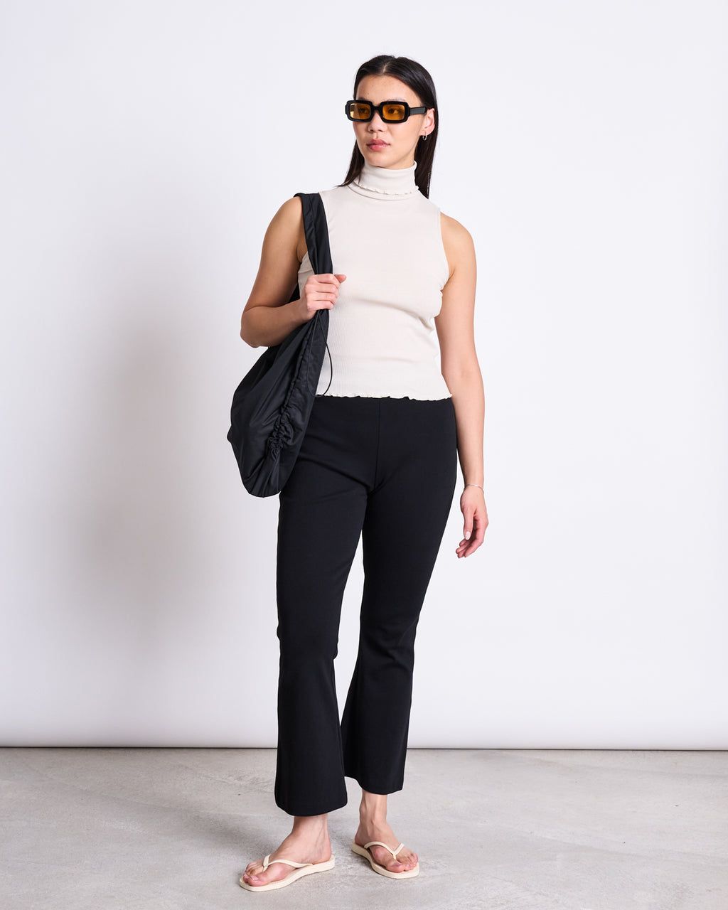A woman in a sleeveless beige turtleneck, jan-n-june FLARED PANTS MIOSA BLACK GOTS with an elastic waistband, flip-flops, and large sunglasses stands against a white wall holding a large black shoulder bag.