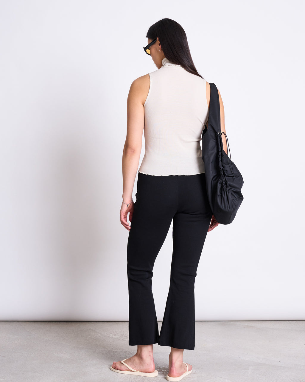 A woman with long dark hair, sunglasses, a sleeveless beige top, jan-n-june FLARED PANTS MIOSA BLACK GOTS with an elastic waistband, and flip-flops stands facing away from the camera with a large black shoulder bag against a white background.