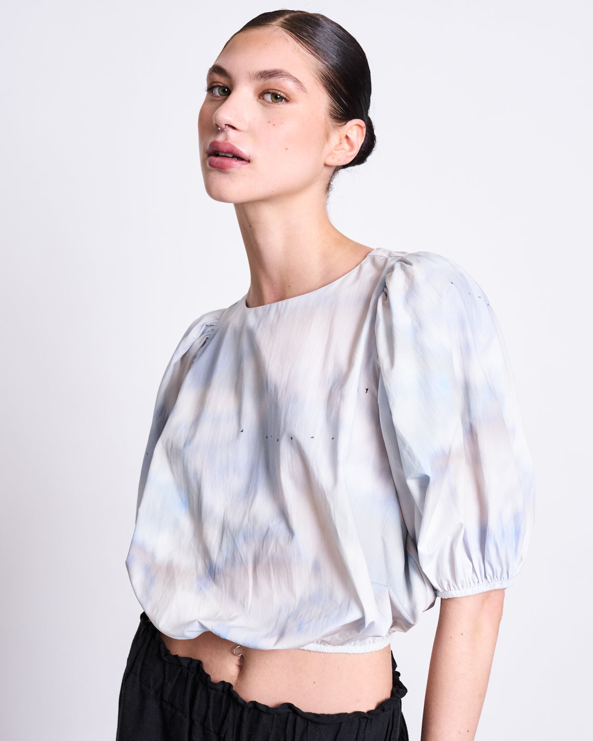 A woman with dark hair in a sleek bun poses confidently against a plain background, wearing the jan-n-june BLOUSE LAIDE CLOUD PRINT with puffy sleeves and black pants.