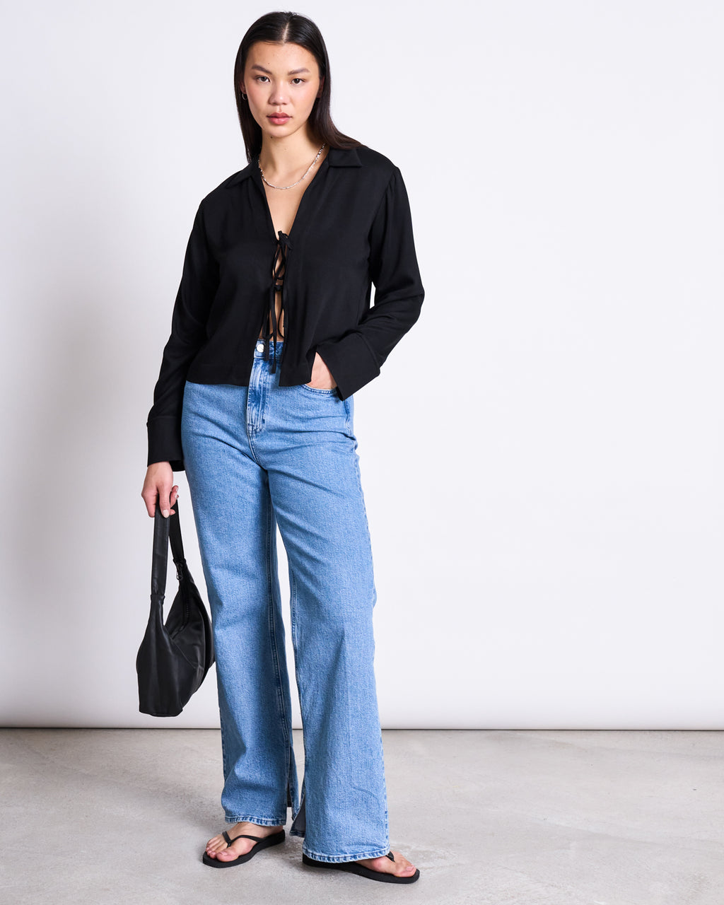 A woman stands against a plain white wall wearing the jan-n-june TIE BLOUSE KALUZ BLACK, light blue wide-leg jeans, black flip-flops, and holding a black handbag.
