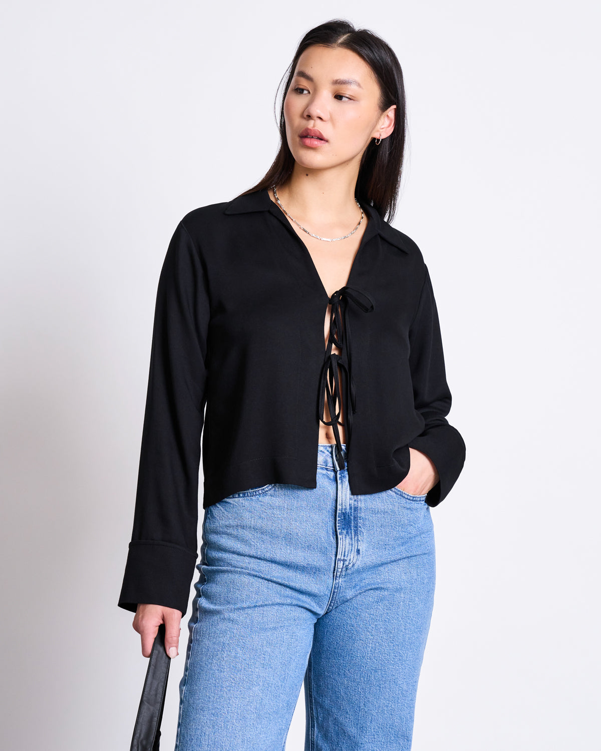 A woman with long dark hair stands against a white background wearing the jan-n-june TIE BLOUSE KALUZ BLACK and light blue high-waisted jeans, looking slightly to the side and holding a black bag.
