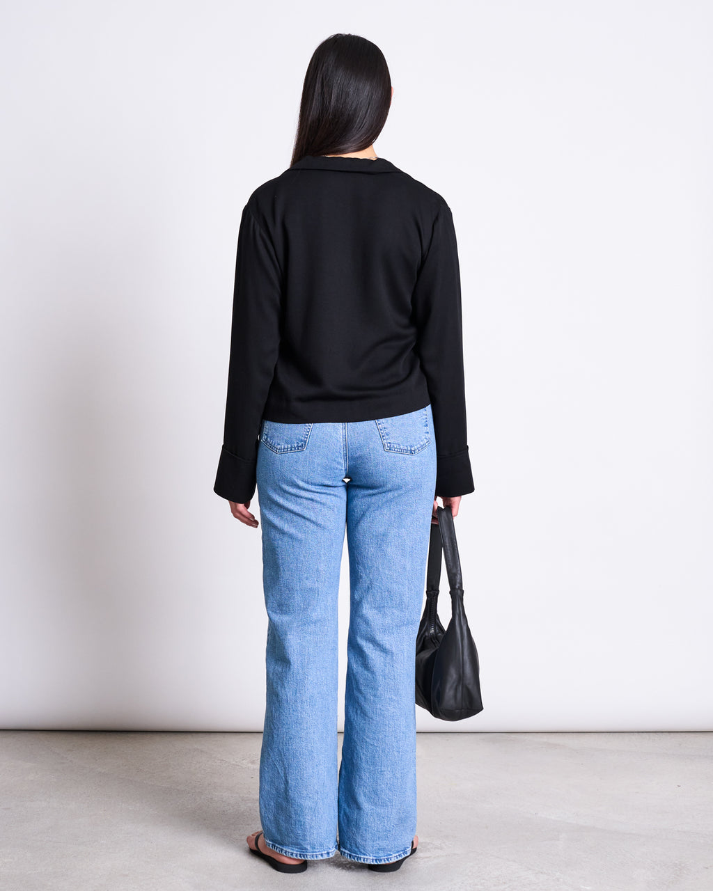 A woman faces away from the camera wearing the jan-n-june TIE BLOUSE KALUZ BLACK, light blue jeans, and black sandals. She holds a black handbag in her left hand against a plain white background.