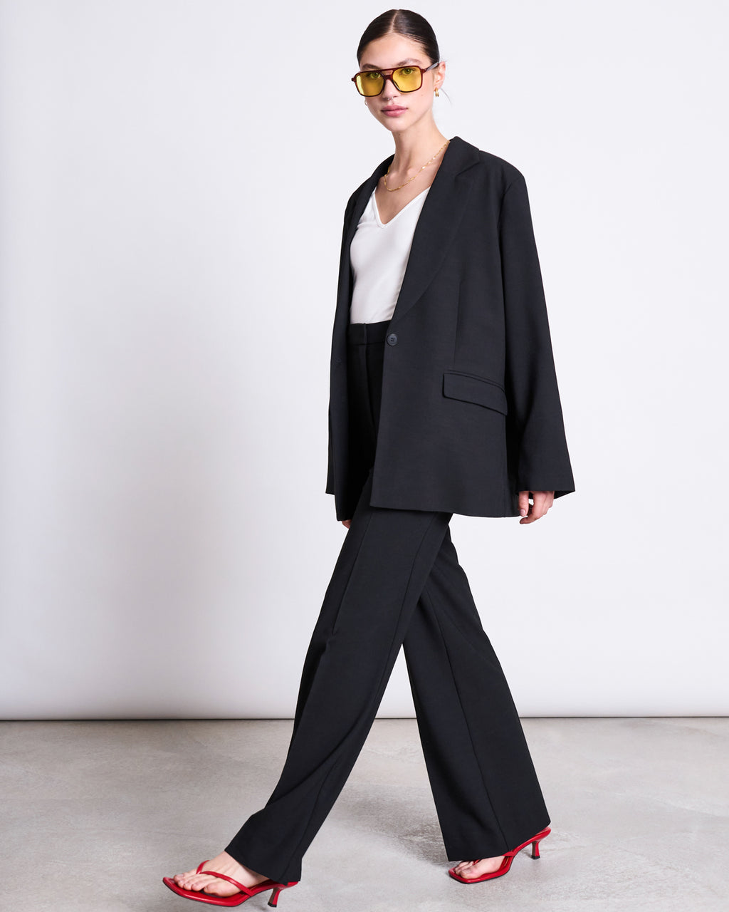 A woman confidently walks against a white background wearing jan-n-junes ECOVERO™ WIDE PANTS CORALIE BLACK, paired with a white top, yellow-tinted sunglasses, and red heeled sandals.