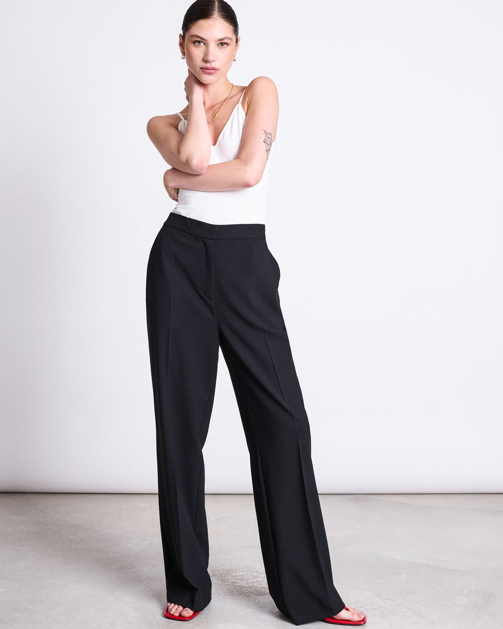 A woman poses against a white background wearing jan-n-june’s ECOVERO™ WIDE PANTS CORALIE BLACK, paired with a white sleeveless top and red open-toe heels. Her hair is pulled back, and she thoughtfully rests her arm on her chin.