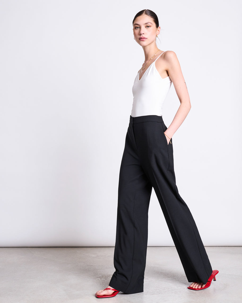 A woman stands against a plain background wearing jan-n-junes ECOVERO™ WIDE PANTS CORALIE BLACK, a white sleeveless top, and red heeled sandals. Hands in pockets, she looks confidently at the camera.