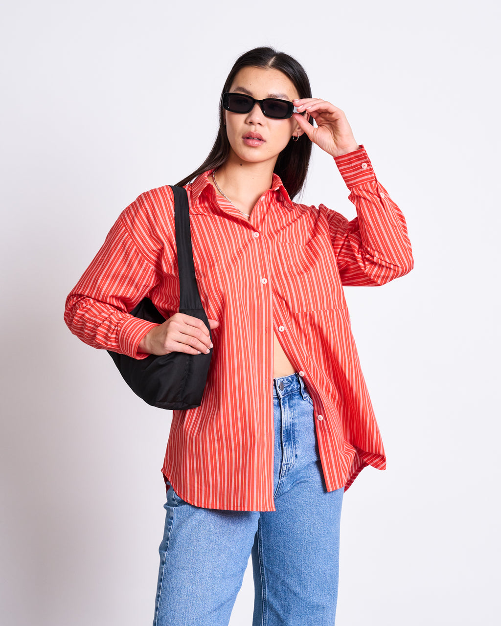 A woman models the jan-n-june OVERSIZED SHIRT MAMRO CHERRY RED WHITE STRIPED GOTS, styled with blue jeans, black sunglasses, and a black shoulder bag against a plain white background.