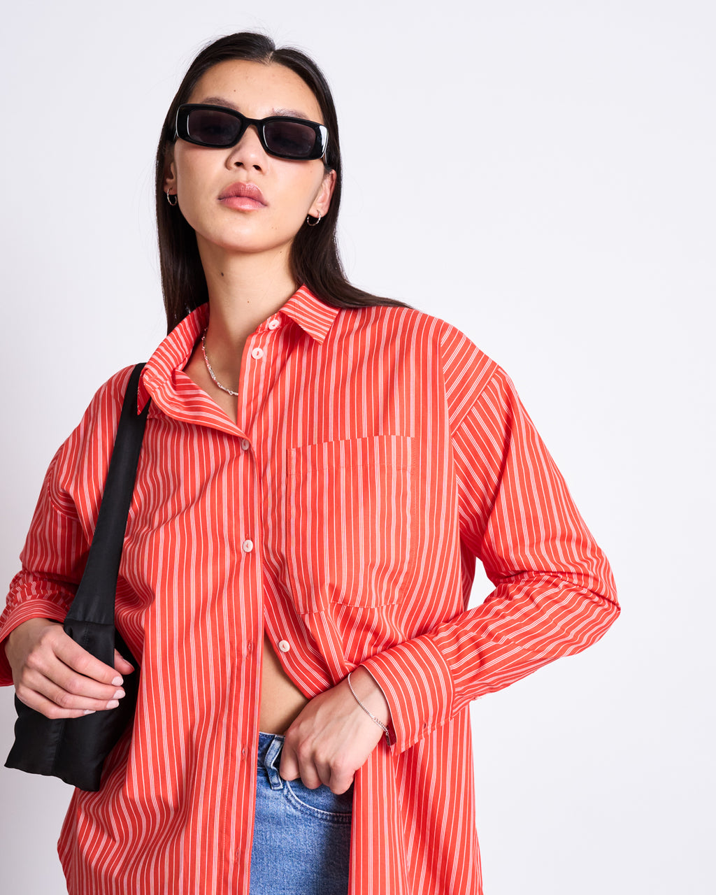 A woman models the jan-n-june OVERSIZED SHIRT MAMRO CHERRY RED WHITE STRIPED GOTS, paired with blue jeans and black sunglasses, posing confidently with a black bag against a plain white background.