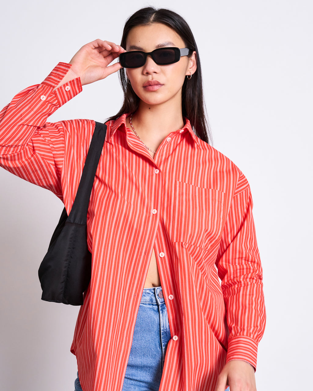 A woman wearing the jan-n-june OVERSIZED SHIRT MAMRO CHERRY RED WHITE STRIPED GOTS, blue jeans, and black sunglasses poses with a black shoulder bag against a plain white background. She has straight dark hair and touches her glasses.