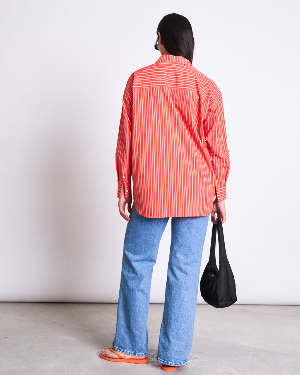 A person stands facing away, wearing the jan-n-june OVERSIZED SHIRT MAMRO CHERRY RED WHITE STRIPED GOTS, light blue jeans, and orange sandals. They hold a black bag in their left hand against a plain white background.
