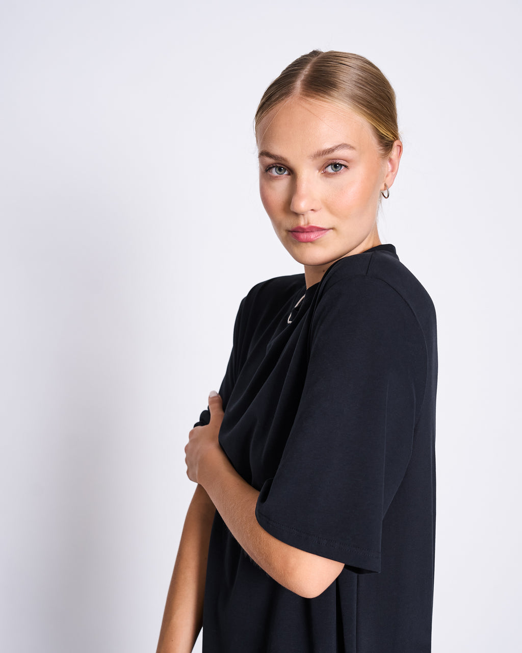 A woman with blonde hair in a low bun wears the jan-n-june SHIRT DRESS CROVIE BLACK made from recycled cotton. She stands against a white background, looking at the camera with a neutral expression and one arm crossed over her chest.