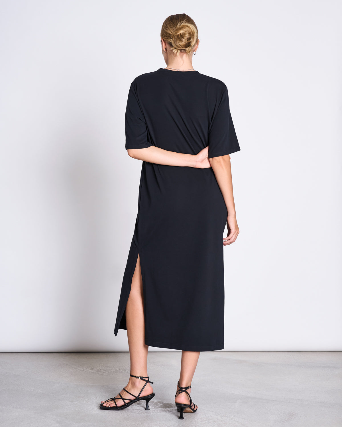 A woman faces away, wearing the jan-n-june SHIRT DRESS CROVIE BLACK with short sleeves and a side slit, paired with black strappy heeled sandals. Her hair is in a neat bun, one arm behind her back, against a plain background.